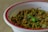 A white bowl with a red rim containing a dish made of cooked chickpeas. The chickpeas are seasoned and garnished with fresh cilantro leaves and spices. The dish appears to have green chili and onions mixed in for flavor.