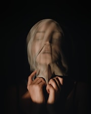 A person is shrouded in translucent fabric, with soft shadows and highlights creating a mysterious and ethereal effect. The facial features are slightly obscured, conveying a sense of enigma or introspection.