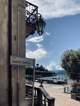 A scenic street view with a sign labeled 'via isola bella' mounted on a building. An ornate metal lamp hangs from an upper balcony. Below is a railing and a cobblestone road with trees lining the waterfront. Boats are visible on the water under a partly cloudy sky, with mountains in the distance.