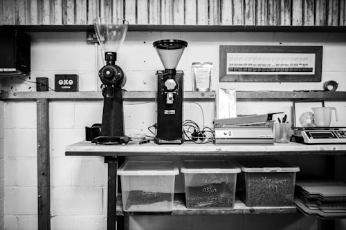 A black and white photo of a workspace with two coffee grinders placed on a wooden counter. Below the counter are transparent containers labeled with coffee beans inside. The wall has various items including a framed chart and shelves with bags. Various equipment like scales and a sealing machine are present along with a cup and writing tools.