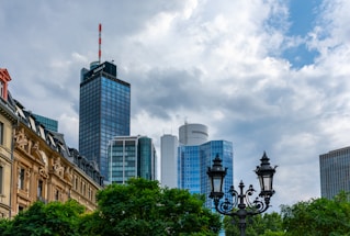 A cityscape featuring modern skyscrapers with a prominent black glass building labeled 'Helaba' and surrounding high-rise structures with reflective windows. In the foreground, there is an ornate street lamp with intricate black metalwork. The sky is overcast with a mixture of clouds and blue patches. Traditional European architecture is visible on the left side, with detailed facades and roof designs. Lush green trees border the lower section of the image.
