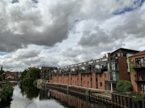 A row of modern apartment buildings with large windows line the edge of a calm river. A series of gray clouds fill the sky, adding a dramatic backdrop. Trees and greenery are visible along the riverbank and on balconies. The river reflects the buildings and sky, creating a tranquil scene.