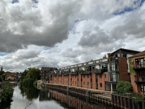 A row of modern apartment buildings with large windows line the edge of a calm river. A series of gray clouds fill the sky, adding a dramatic backdrop. Trees and greenery are visible along the riverbank and on balconies. The river reflects the buildings and sky, creating a tranquil scene.