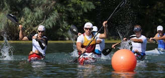 A group of athletes in kayaks are competing in a race on a body of water. They are paddling vigorously, creating splashes around them. The foreground features an orange buoy, while the background consists of lush greenery.