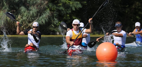 A group of athletes in kayaks are competing in a race on a body of water. They are paddling vigorously, creating splashes around them. The foreground features an orange buoy, while the background consists of lush greenery.