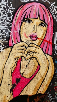 A vibrant mural depicting a stylized woman with pink hair and bright pink lips, holding her hands in front of her face. The background features a graffiti-covered wall with various tags and scribbles. The artwork's bold lines and vivid colors create a striking visual impact.
