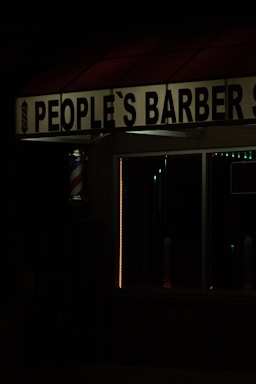 The image features a dimly lit barbershop with a sign reading 'People's Barber Shop'. A classic barber pole with red, white, and blue stripes is visible next to the entrance. The dark ambiance suggests it may be nighttime.