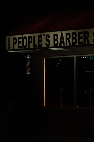 The image features a dimly lit barbershop with a sign reading 'People's Barber Shop'. A classic barber pole with red, white, and blue stripes is visible next to the entrance. The dark ambiance suggests it may be nighttime.