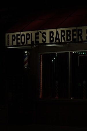 The image features a dimly lit barbershop with a sign reading 'People's Barber Shop'. A classic barber pole with red, white, and blue stripes is visible next to the entrance. The dark ambiance suggests it may be nighttime.