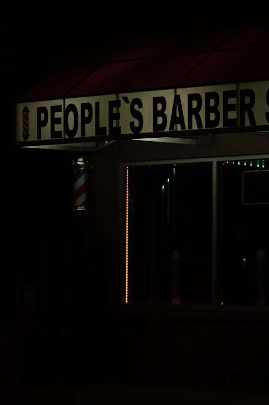 The image features a dimly lit barbershop with a sign reading 'People's Barber Shop'. A classic barber pole with red, white, and blue stripes is visible next to the entrance. The dark ambiance suggests it may be nighttime.