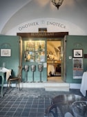 A quaint restaurant entrance with arched ceilings and a sign reading 'Onothek Enoteca' above the door. The door is open, revealing a warmly lit interior with tables set for dining. Wine bottles are displayed in a window to the left of the entrance. Three metal ice buckets are mounted on stands just outside the door. There is a cobblestone floor in the foreground and green wooden panels on the walls. A vintage-style lantern hangs from the ceiling.