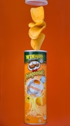 A canister of Pringles with paprika flavor is displayed against an orange background. The canister features a design with a football theme, including a soccer ball image. Multiple chips are seen flowing out of the top of the open canister.