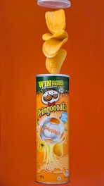 A canister of Pringles with paprika flavor is displayed against an orange background. The canister features a design with a football theme, including a soccer ball image. Multiple chips are seen flowing out of the top of the open canister.