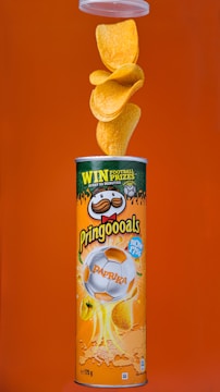 A canister of Pringles with paprika flavor is displayed against an orange background. The canister features a design with a football theme, including a soccer ball image. Multiple chips are seen flowing out of the top of the open canister.
