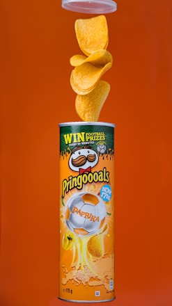 A canister of Pringles with paprika flavor is displayed against an orange background. The canister features a design with a football theme, including a soccer ball image. Multiple chips are seen flowing out of the top of the open canister.