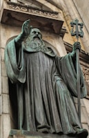 A large bronze statue of a robed figure holding a staff with a cross stands prominently on a stone facade. The figure has a long beard and is positioned beneath an arch with the inscription 'SÃO BENTO'. The statue exudes a sense of solemnity and authority, with intricate details in the robe and facial expressions.