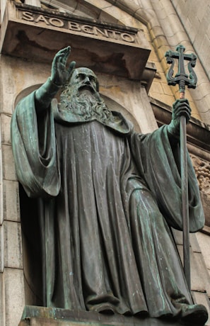 A large bronze statue of a robed figure holding a staff with a cross stands prominently on a stone facade. The figure has a long beard and is positioned beneath an arch with the inscription 'SÃO BENTO'. The statue exudes a sense of solemnity and authority, with intricate details in the robe and facial expressions.