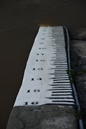 A vertical concrete structure marked with large black numbers and lines measuring water levels. It is placed next to a body of water, possibly a river, showing various measurements from 9 to 16. The background includes dark, flowing water and part of a rocky surface.