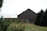 A rustic wooden barn stands in a grassy field surrounded by a well-maintained garden with plants and a few visible fruits. Tall evergreen trees border the setting, and a clear blue sky provides a serene backdrop. A vintage metal lamp post extends above the barn roof, adding to the rural charm.