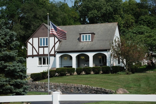 A two-story house with Tudor-style architecture, featuring a prominent front porch with arches. An American flag is displayed on a flagpole in the yard. The house is set amidst a well-maintained lawn and a variety of trees, providing a serene and picturesque environment.