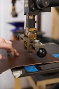 A hand operates a machine press on a piece of brown leather. The machine appears industrial with metallic components and a handle. The background shows blurred elements, suggesting a workshop or factory setting.