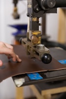 A hand operates a machine press on a piece of brown leather. The machine appears industrial with metallic components and a handle. The background shows blurred elements, suggesting a workshop or factory setting.