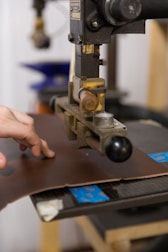 A hand operates a machine press on a piece of brown leather. The machine appears industrial with metallic components and a handle. The background shows blurred elements, suggesting a workshop or factory setting.