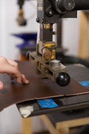A hand operates a machine press on a piece of brown leather. The machine appears industrial with metallic components and a handle. The background shows blurred elements, suggesting a workshop or factory setting.