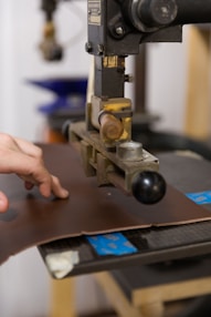 A hand operates a machine press on a piece of brown leather. The machine appears industrial with metallic components and a handle. The background shows blurred elements, suggesting a workshop or factory setting.