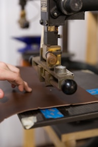A hand operates a machine press on a piece of brown leather. The machine appears industrial with metallic components and a handle. The background shows blurred elements, suggesting a workshop or factory setting.