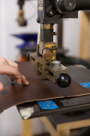 A hand operates a machine press on a piece of brown leather. The machine appears industrial with metallic components and a handle. The background shows blurred elements, suggesting a workshop or factory setting.