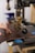 A hand operates a machine press on a piece of brown leather. The machine appears industrial with metallic components and a handle. The background shows blurred elements, suggesting a workshop or factory setting.