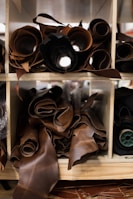 Multiple rolled-up sheets of leather are organized on wooden shelves. The leather is primarily dark brown, with some variably textured surfaces. The rolls are neatly arranged, showcasing a variety of leather types and thicknesses.