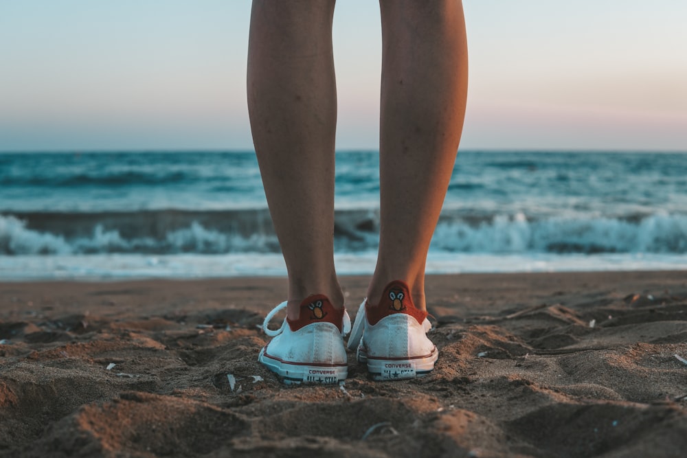 person wearing white low top sneakers standing on brown sand near sea during daytime