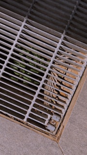 A metal grate covers an opening on a concrete surface. Sunlight casts shadow patterns of the grate bars onto the surrounding area. There is some dirt and possible plant life visible beneath the grate.