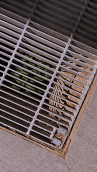 A metal grate covers an opening on a concrete surface. Sunlight casts shadow patterns of the grate bars onto the surrounding area. There is some dirt and possible plant life visible beneath the grate.