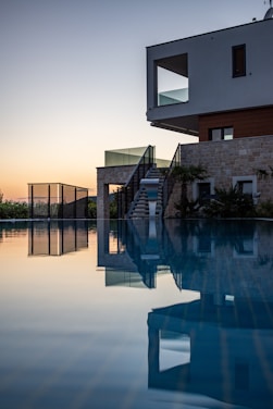 A modern house with minimalist architecture is reflected in a calm swimming pool at dusk. The building features stone and wood cladding with large windows, and a set of stairs leading to the pool area. The serene atmosphere is accentuated by the soft glow of the setting sun in the background.