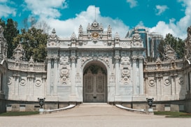An ornate and grand entrance gate with intricate carvings and decorative elements dominates the scene. The structure is symmetrical with elaborate stonework, showcasing a blend of architectural styles. In the background, lush trees and modern buildings are partially visible, creating a contrast between the historical and contemporary. The sky is a bright blue with scattered clouds.