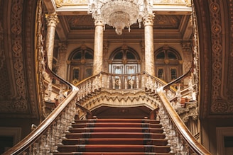 A grand, opulent interior featuring a sweeping double staircase with ornate railings and rich carpeting leads up to a set of grand doors. Intricate architectural details such as carved columns, adorned moldings, and decorative elements are visible throughout. A large, elaborate chandelier hangs from the ceiling, enhancing the luxurious atmosphere.