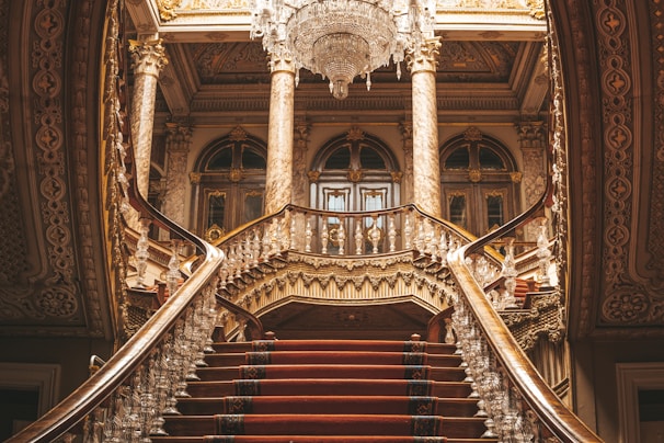 A grand, opulent interior featuring a sweeping double staircase with ornate railings and rich carpeting leads up to a set of grand doors. Intricate architectural details such as carved columns, adorned moldings, and decorative elements are visible throughout. A large, elaborate chandelier hangs from the ceiling, enhancing the luxurious atmosphere.