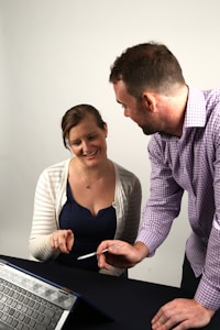 A woman and a man are engaged in a collaborative discussion over a tablet placed on a table. The woman is seated, gesturing towards the tablet, while the man is standing and holding a stylus, seemingly explaining or pointing something out on the screen.