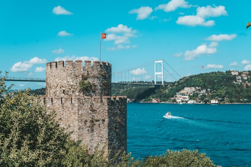 A historic stone tower is in the foreground, featuring a Turkish flag on top. The tower is surrounded by lush greenery. In the background, large bridge spans across a wide, blue river with a small boat traveling along it. A hilly landscape with residential buildings is visible on the other side of the river under a bright blue sky scattered with fluffy clouds.