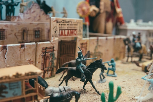 A detailed miniature diorama representing a historical battle scene. Tiny figurines of soldiers, some on horseback, are positioned in front of a small replica of a fort with 'Alamo' written on it. The scene is set in a desert-like environment with scattered cacti, and the overall arrangement suggests a historical re-enactment or display.