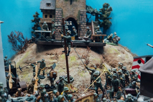 A scene featuring miniature soldier figurines engaged in various activities on a hilly terrain. At the center, a figure is climbing a pole with bells, while others are gathered around appearing to be performing tasks or in combat. In the background, there is a building that resembles a medieval castle or fort, surrounded by miniature trees and other military personnel. Some soldiers are holding tools and flags.