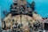 A scene featuring miniature soldier figurines engaged in various activities on a hilly terrain. At the center, a figure is climbing a pole with bells, while others are gathered around appearing to be performing tasks or in combat. In the background, there is a building that resembles a medieval castle or fort, surrounded by miniature trees and other military personnel. Some soldiers are holding tools and flags.