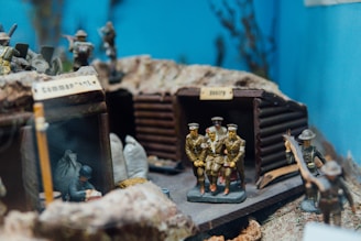 A diorama depicting a wartime scene with miniature soldiers. Several figures are positioned in front of wooden structures resembling trenches or bunkers, one labeled 'Command', and others marked 'Entry'. The soldiers are dressed in uniforms from the early 20th century, with some carrying rifles and one figure appearing to be carrying a wounded comrade. Sandbags and other military accessories are present, suggesting a battlefield setting. The background is a blue sky with indistinct vegetation.