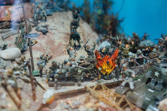 A diorama depicts a wartime scene with toy soldiers in combat. The landscape includes a road sign pointing to Berlin, smoke and fire effects simulating an explosion, and various soldiers in different poses, some appearing to be shooting, crouched or lying down, possibly injured. Vegetation and a tank add to the battlefield ambiance.