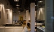 A modern furniture showroom with several sofas and beds displayed. The interior is illuminated with ceiling lights and features white brick columns. A person is seen sitting on a chair in the background.