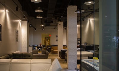 A modern furniture showroom with several sofas and beds displayed. The interior is illuminated with ceiling lights and features white brick columns. A person is seen sitting on a chair in the background.
