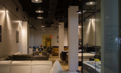 A modern furniture showroom with several sofas and beds displayed. The interior is illuminated with ceiling lights and features white brick columns. A person is seen sitting on a chair in the background.